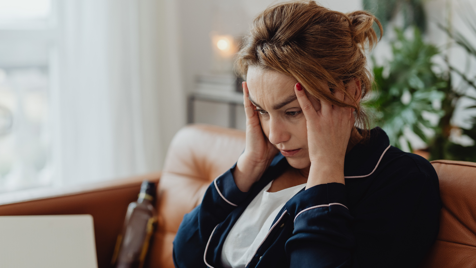 A woman sitting indoors, covering her had with her hands in a gesture that might express stress, sadness, or fatigue, seeking a moment of alignment.