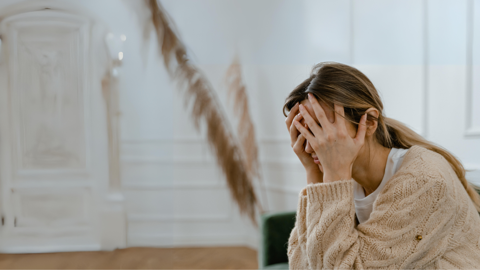 A woman in a cozy sweater sitting indoors, covering her face with her hands in a gesture that might express stress, sadness, or fatigue, seeking a moment of alignment.